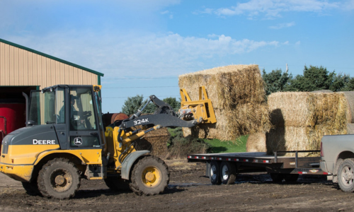 A forklift on a farm loading square bales on a flat bed.